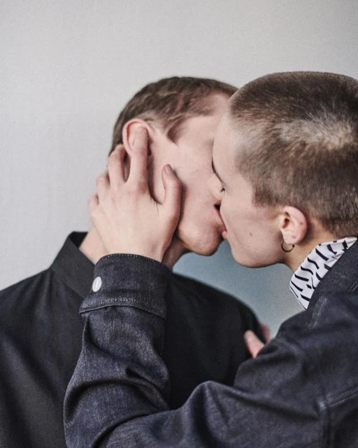 Close-up portrait of two young adult fair-skinned people kissing, wearing a dark shirt and jean jacket against a grey background.