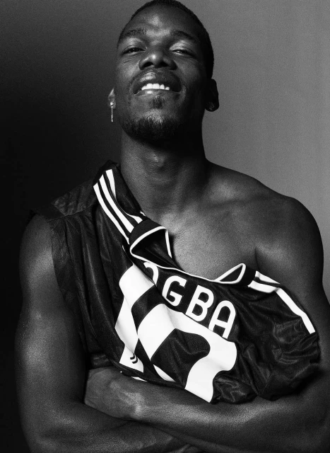 Black and white portrait of bare-chested man in athletic jersey with arms crossed, grinning against a dark gradient backdrop.
