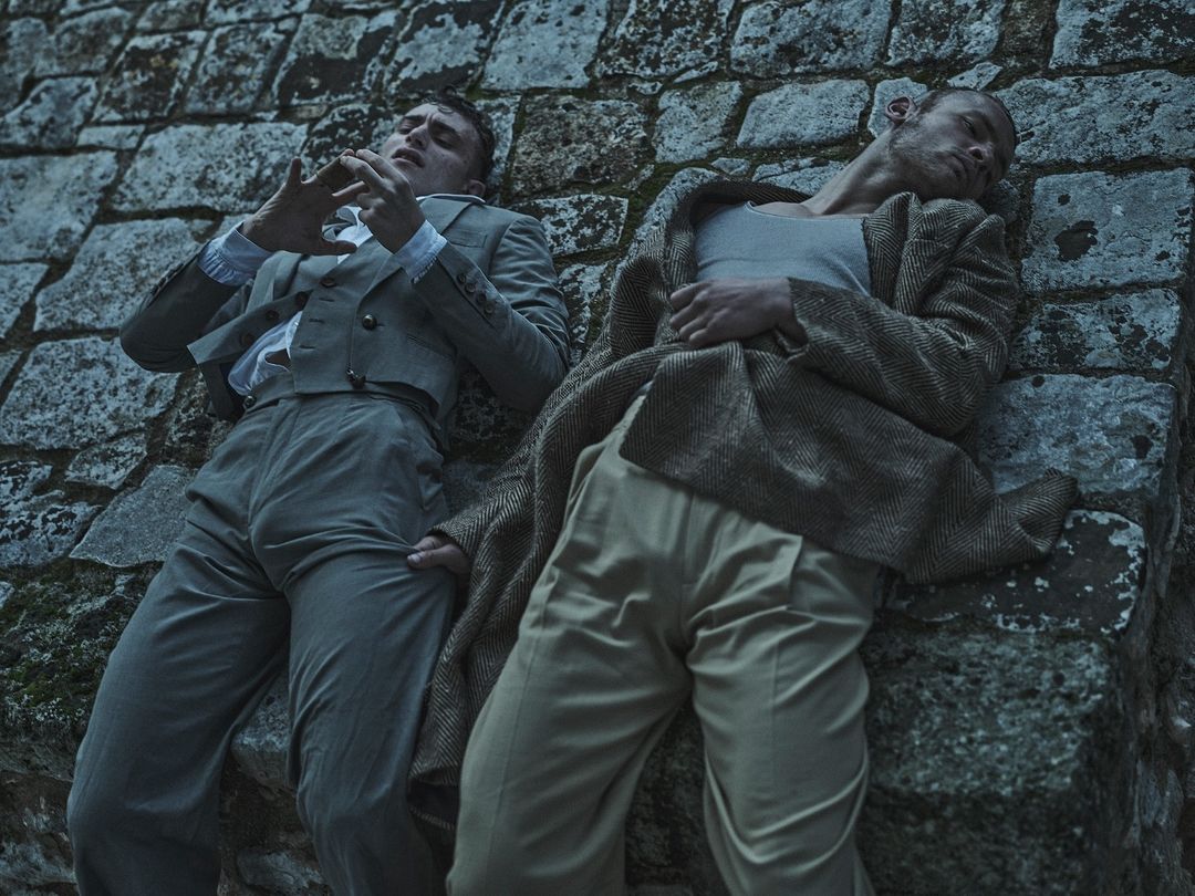 Two men in retro attire recline against a stone structure, one in a suit and the other in a vest and coat, creating a vintage aesthetic.