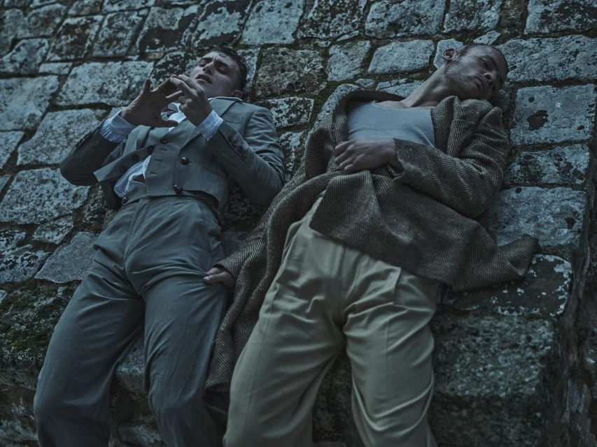 Two men in retro attire recline against a stone structure, one in a suit and the other in a vest and coat, creating a vintage aesthetic.