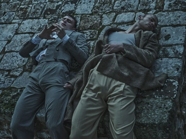Two men in retro attire recline against a stone structure, one in a suit and the other in a vest and coat, creating a vintage aesthetic.