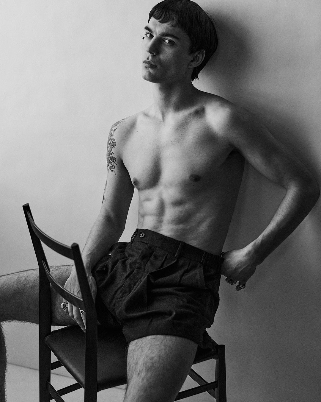 Black and white portrait of a shirtless male model with dark hair, sitting on a chair, looking at the camera in a photo studio.