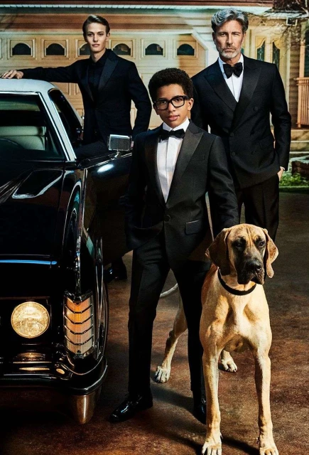 Three men in formal wear pose with a dog and a vintage car in front of a garage, suggesting a formal event or gathering.