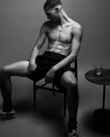 Shirtless man wearing dark shorts sitting on a chair next to a small round table in a studio setting.