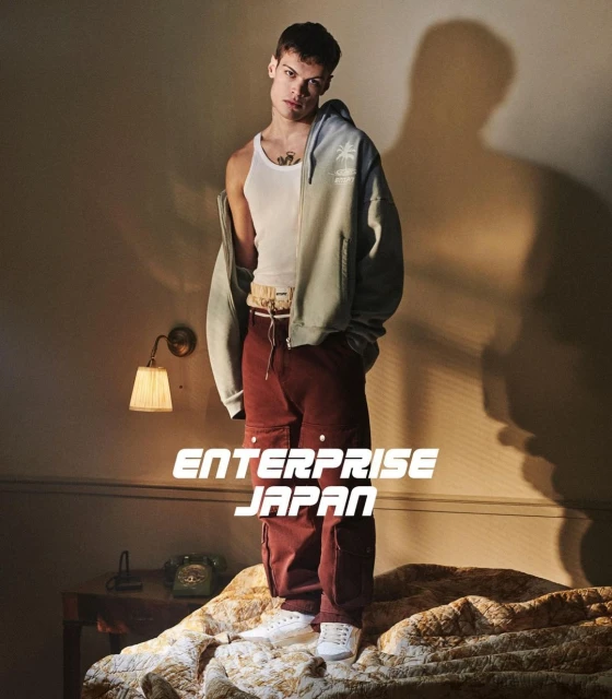 Adult male fashion model poses indoors wearing a white tank top, two-tone hoodie, cargo pants, and sneakers on a bed with patterned bedding.