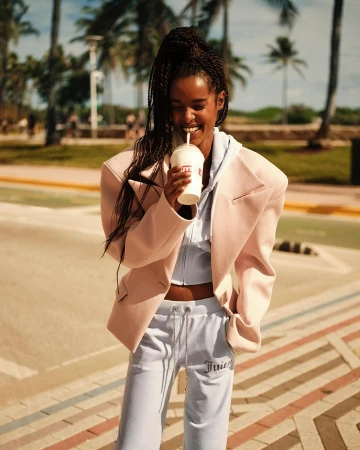A smiling Black woman in a pink blazer and Juicy Couture tracksuit holds a drink in a sidewalk scene with palm trees.