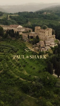 Aerial view of a European-style village with stone buildings and lush green hills in the background, branded with the Paul & Shark logo.