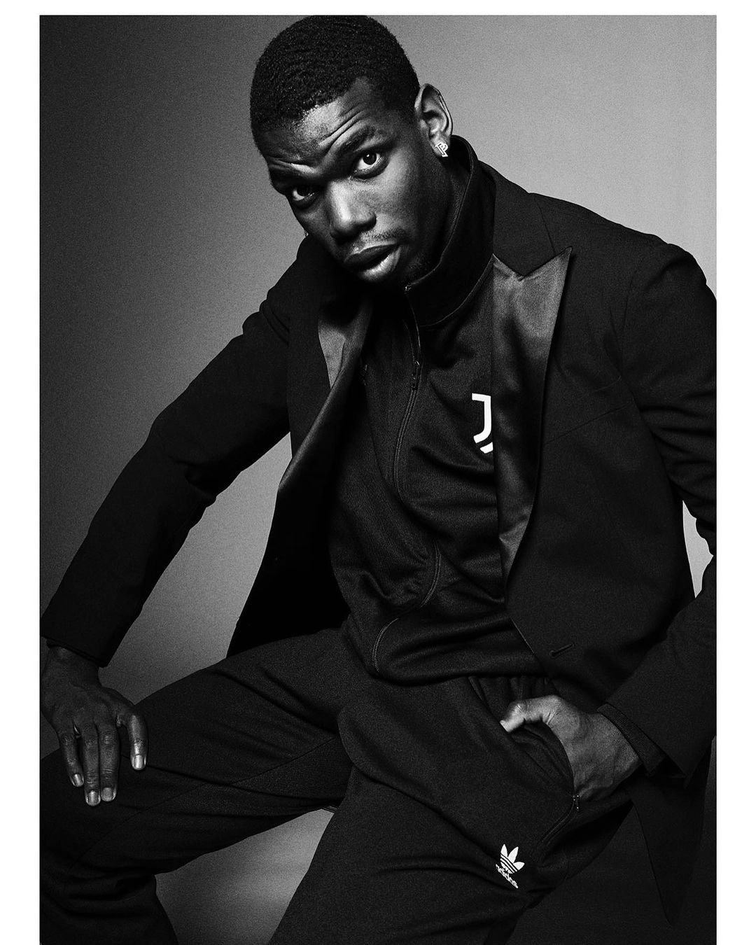 Black and white portrait of an adult male in a blazer over athletic wear sitting in a studio setting, looking at the camera.