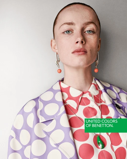 Close-up of a fair-skinned model with short hair wearing a lavender and white polka dot coat, coral earrings, and a red and white polka dot top.