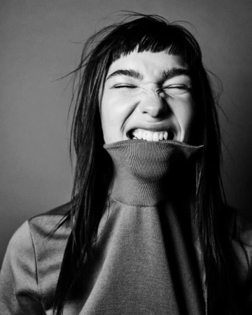 Black and white portrait of a young woman with dark hair and a turtleneck sweater, pulling the collar over her mouth and grinning intensely.