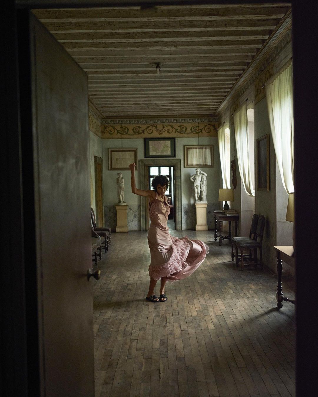 Woman in a pink dress twirls in a hallway lined with marble statues and antique furniture, light streaming through sheer curtains.