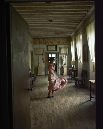 Woman in a pink dress twirls in a hallway lined with marble statues and antique furniture, light streaming through sheer curtains.