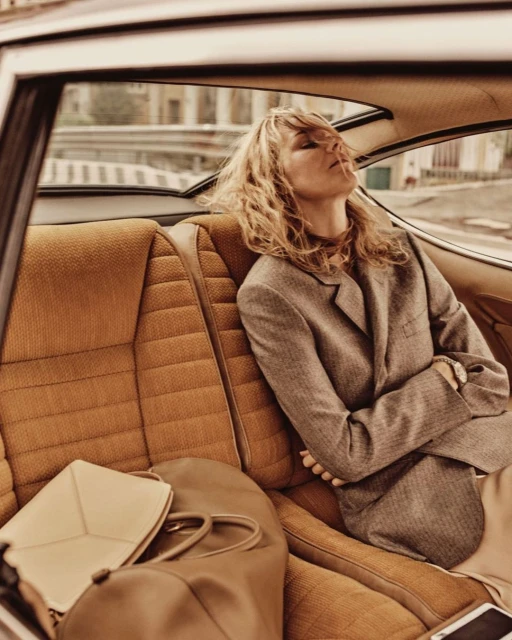 A woman with wavy blond hair wearing a blazer sits in the backseat of a car. The vintage car's brown seats and a leather bag are visible.