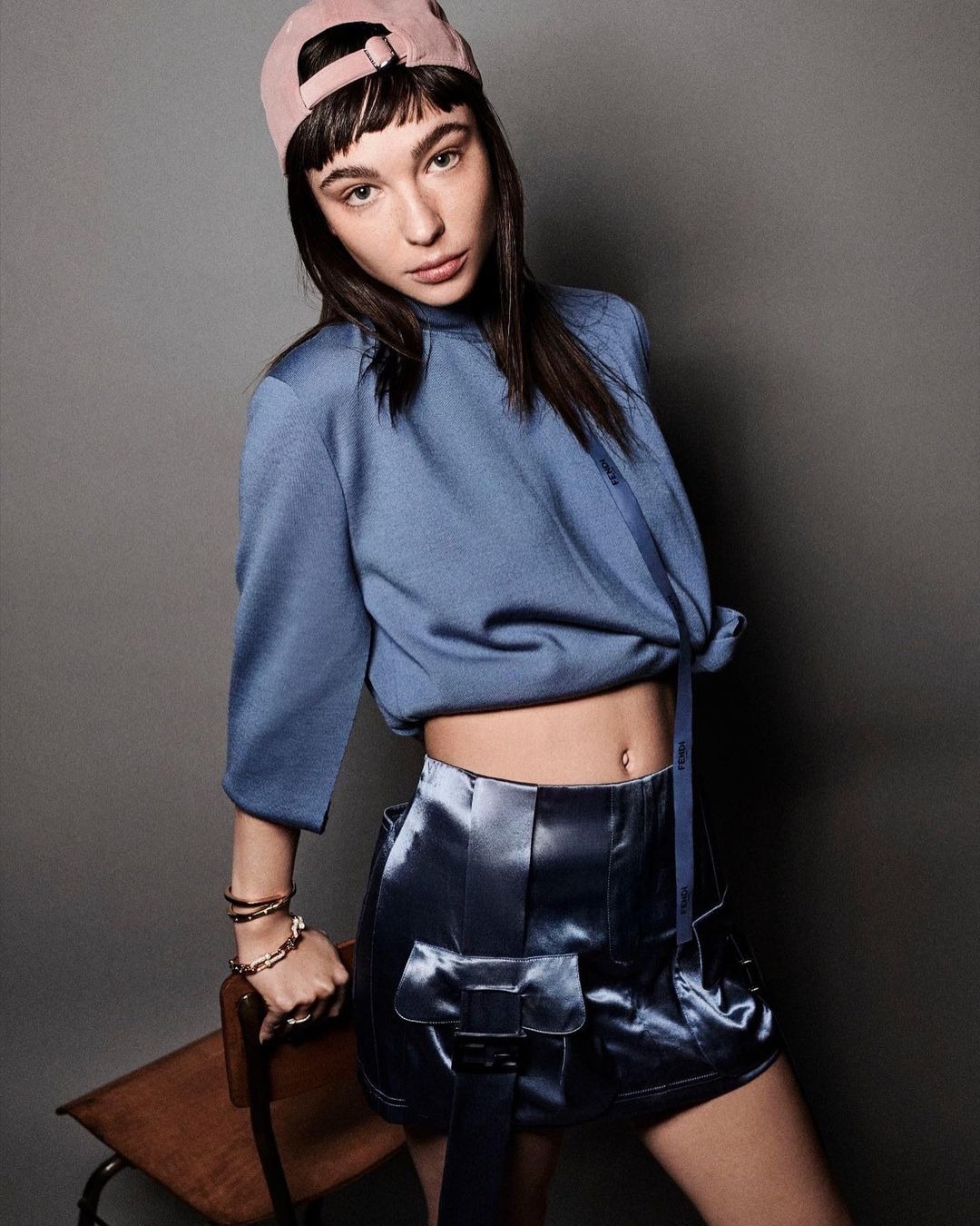 A young adult woman poses indoors wearing a blue top, shiny blue shorts with belt detail, and a pink baseball cap in a studio setting.