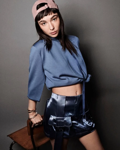 A young adult woman poses indoors wearing a blue top, shiny blue shorts with belt detail, and a pink baseball cap in a studio setting.