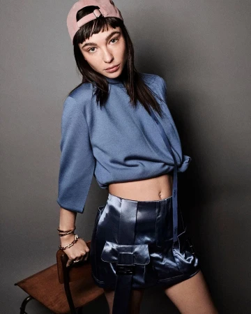 A young adult woman poses indoors wearing a blue top, shiny blue shorts with belt detail, and a pink baseball cap in a studio setting.