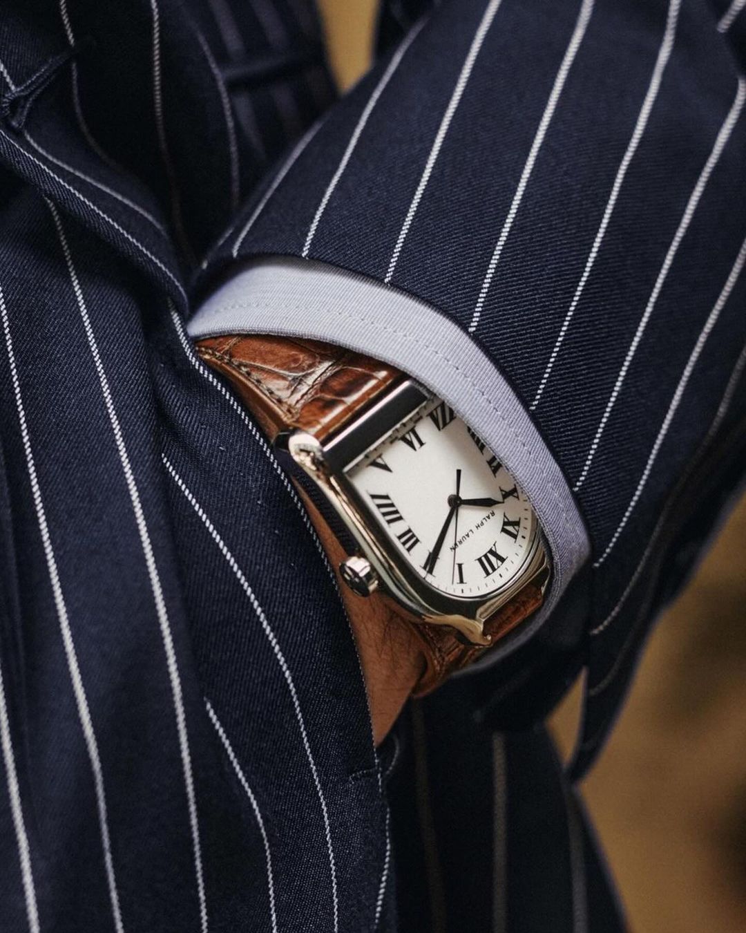 Close-up of a luxury wristwatch on a person's wrist, featuring a tan leather strap and set against a dark pinstripe suit.