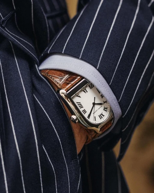 Close-up of a luxury wristwatch on a person's wrist, featuring a tan leather strap and set against a dark pinstripe suit.