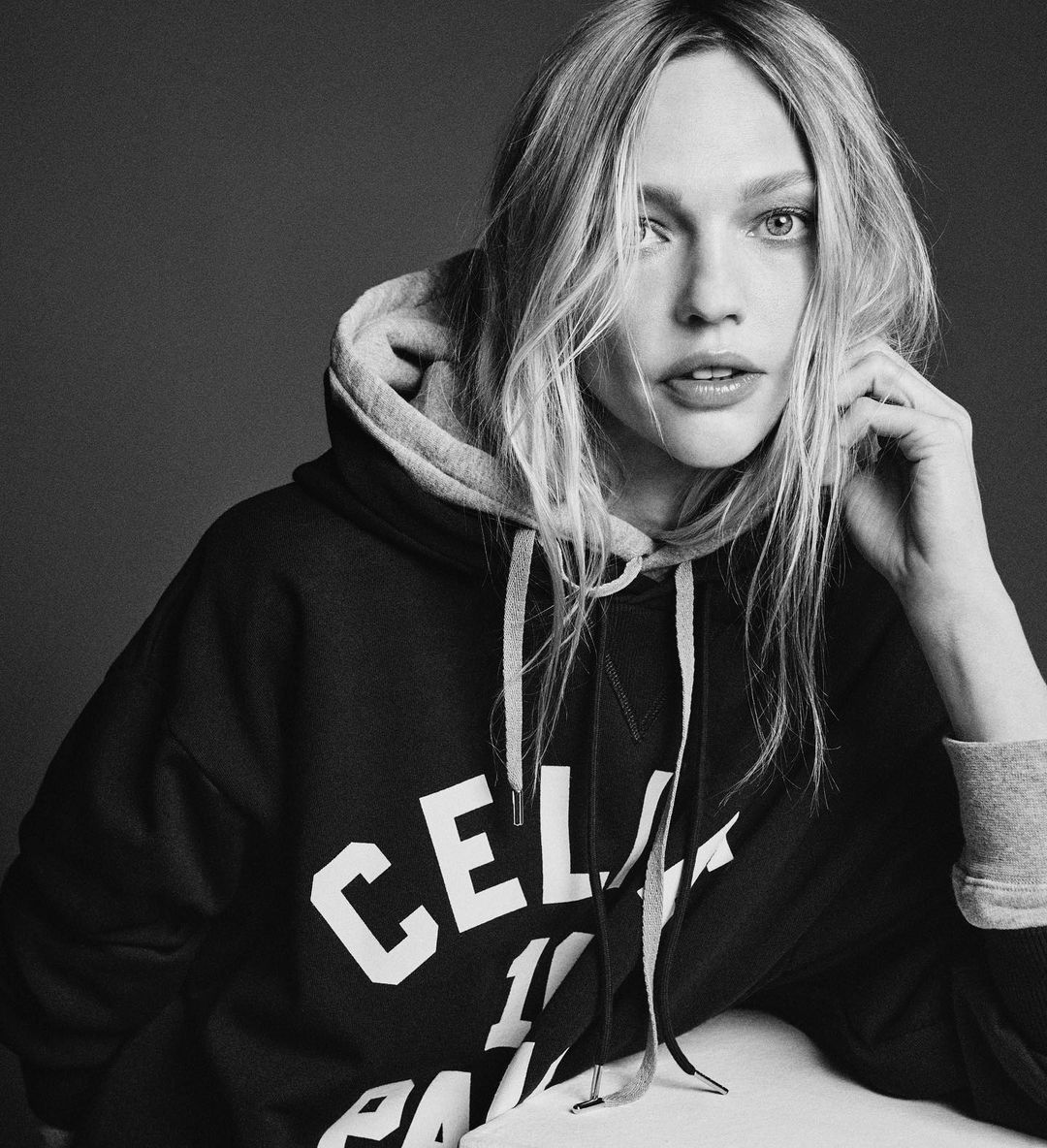 Black and white portrait of a blonde adult female wearing a hooded sweatshirt indoors with a grey background. Close-up view.