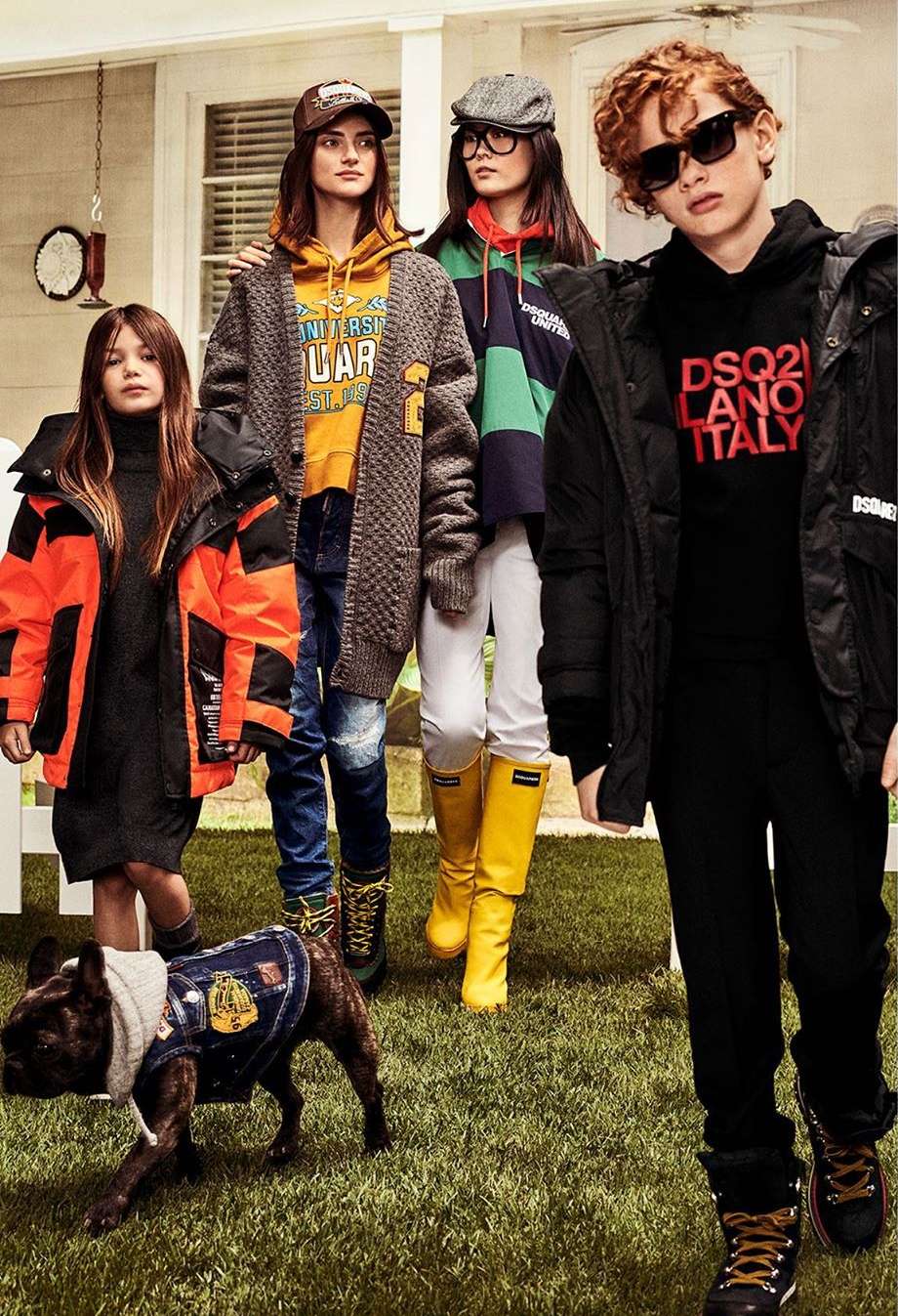 Group of kids in outerwear and boots posing on a grassy lawn in front of a house, including a dog wearing a denim vest and hoodie.