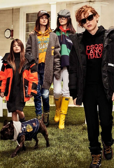 Group of kids in outerwear and boots posing on a grassy lawn in front of a house, including a dog wearing a denim vest and hoodie.