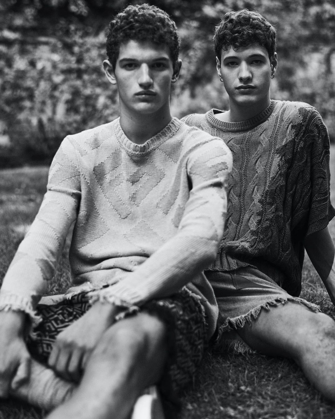 Black and white medium shot of two young adult men in knitted sweaters and shorts sitting on grass outdoors with a stone wall background.