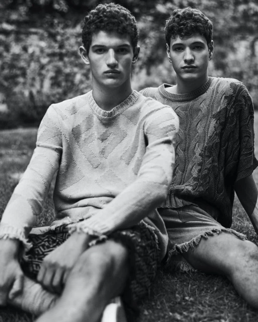 Black and white medium shot of two young adult men in knitted sweaters and shorts sitting on grass outdoors with a stone wall background.