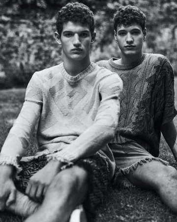Black and white portrait of two male models with curly hair wearing textured knitwear seated outdoors on grass.