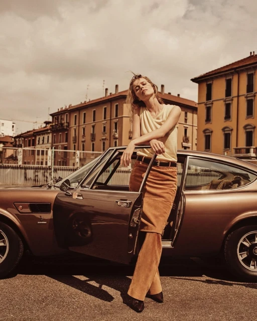 Blonde woman leans on a brown classic car parked on a city street with light brown buildings under a cloudy sky in a fashion-editorial style.