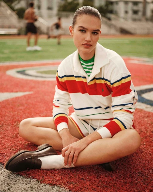 A young woman in a striped rugby shirt sits cross-legged on a sports field with athletic figures visible in the blurred background.