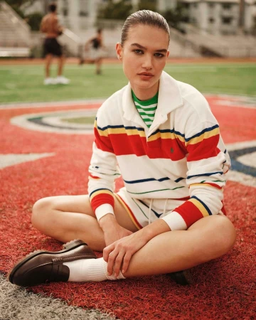 A young woman in a striped rugby shirt sits cross-legged on a sports field with athletic figures visible in the blurred background.