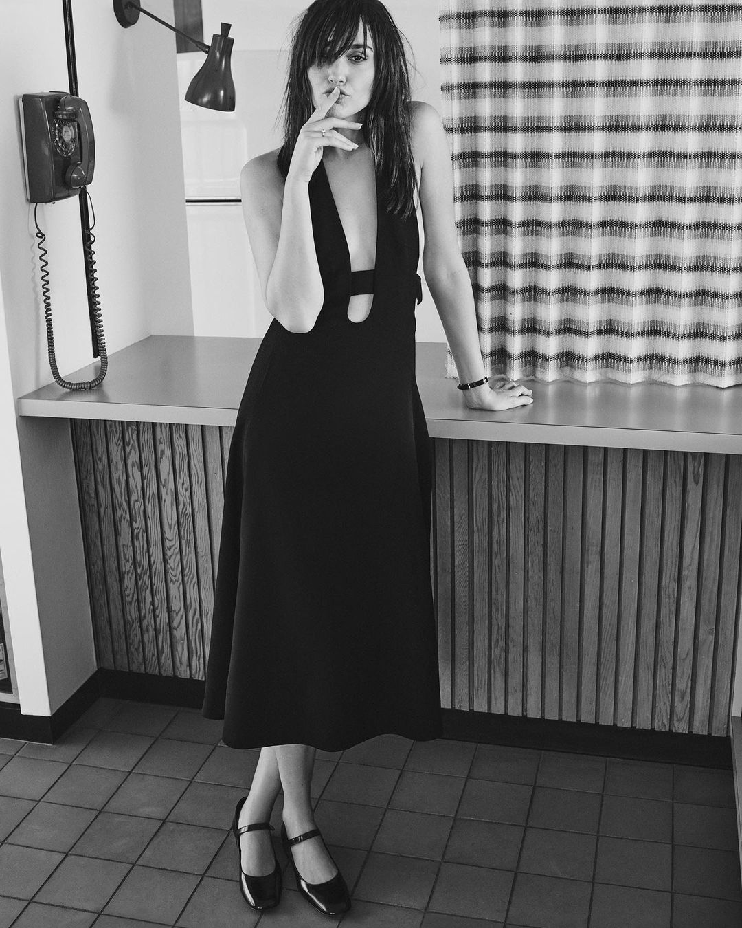 A stylish woman poses indoors wearing a black midi dress and Mary Jane shoes, with a finger to her lips in a shushing gesture.