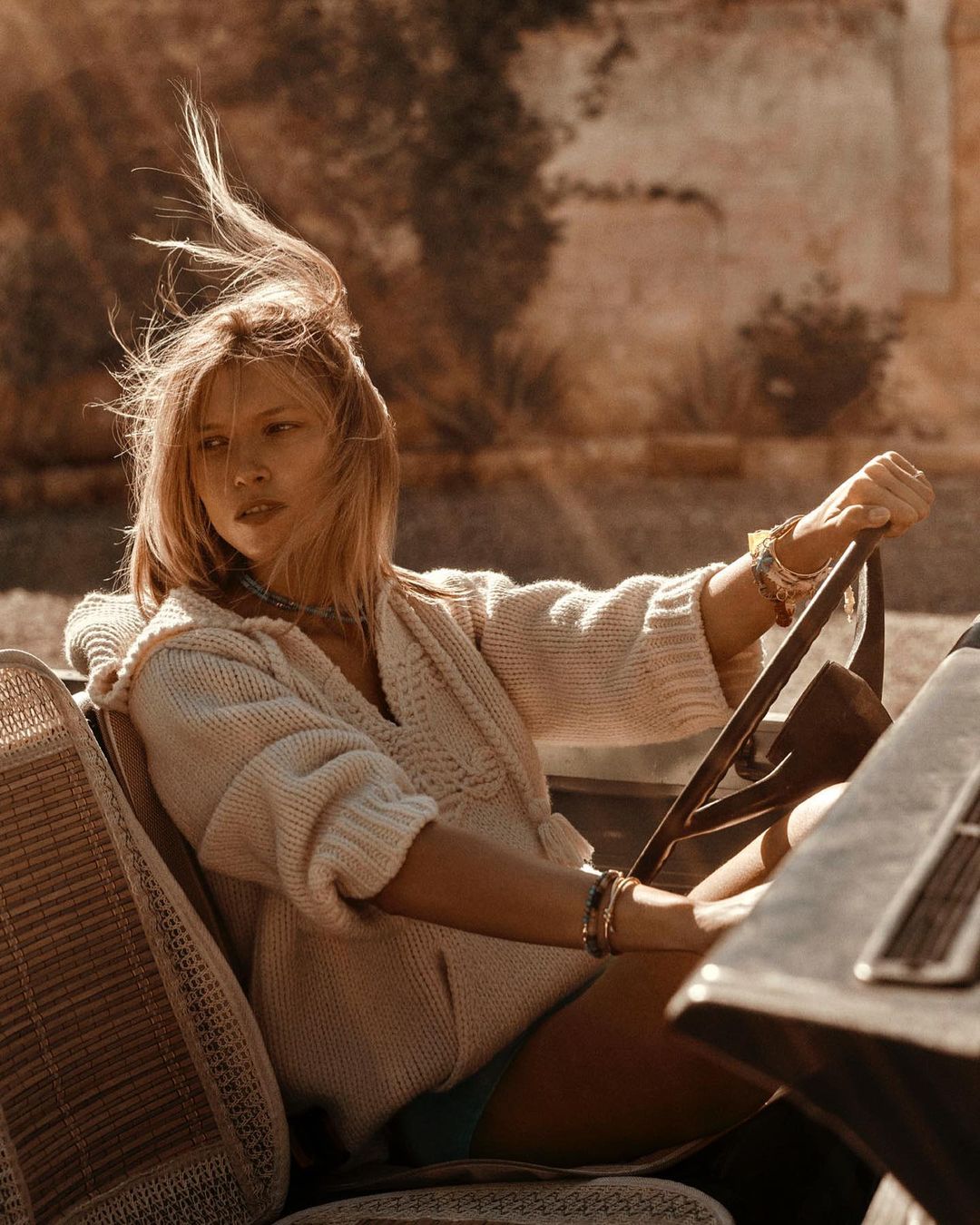 A blonde woman in a hooded sweater drives a vintage car on a sunny day, with her hair blowing in the wind and a light-colored building visible.