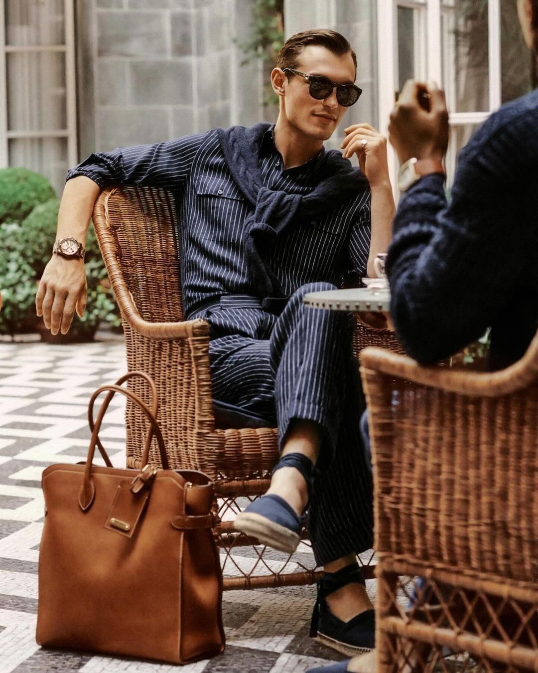 Man in striped pyjama suit seated outside in wicker chair, wearing sunglasses and sweater, holding a cup with a leather tote.