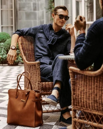 Man in striped pyjama suit seated outside in wicker chair, wearing sunglasses and sweater, holding a cup with a leather tote.