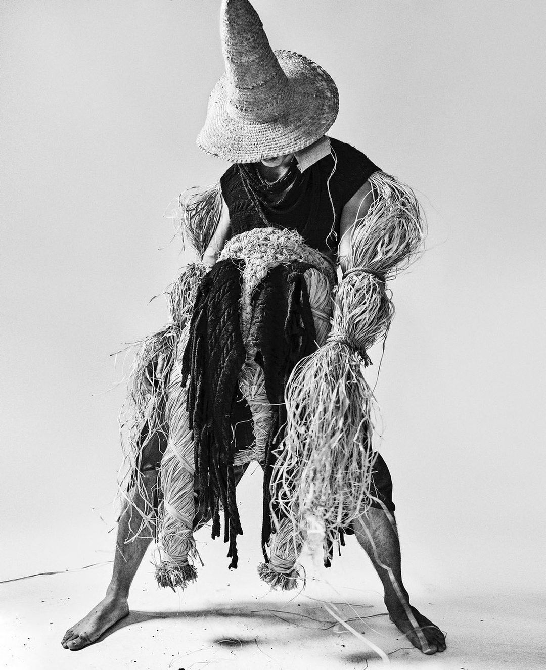 Monochrome studio shot of a person wearing a straw hat and adorned with raffia, posing in front of a white background, artful and textured.