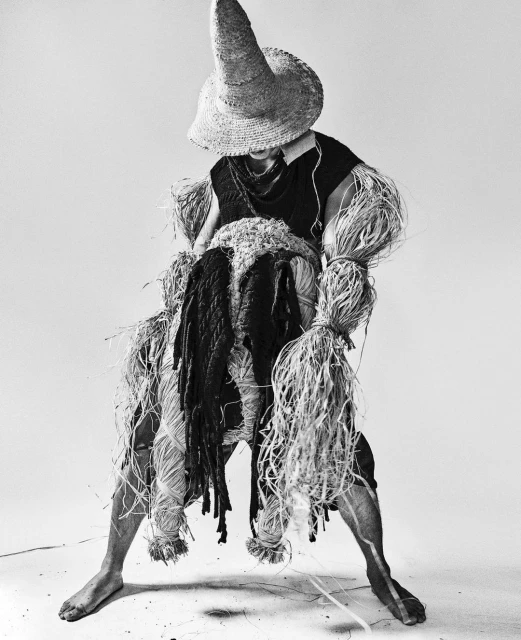 Monochrome studio shot of a person wearing a straw hat and adorned with raffia, posing in front of a white background, artful and textured.