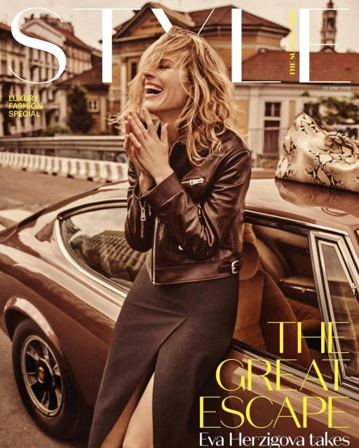 A fashion magazine cover shot featuring a model leaning against a classic car, set against a European street with buildings in the background.