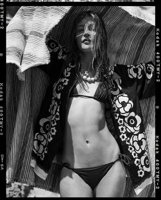 Monochrome portrait of a woman in a bikini and floral jacket, under a striped fabric, captured in a vintage film style with Kodak film borders.