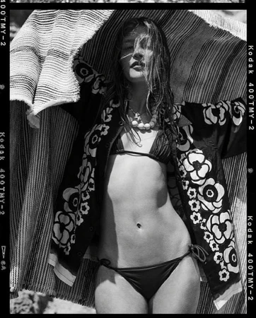 Monochrome portrait of a woman in a bikini and floral jacket, under a striped fabric, captured in a vintage film style with Kodak film borders.