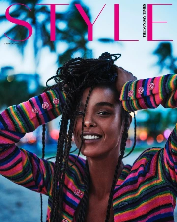 A smiling Black woman with long braided hair wearing a striped colourful cardigan against an outdoor evening backdrop.