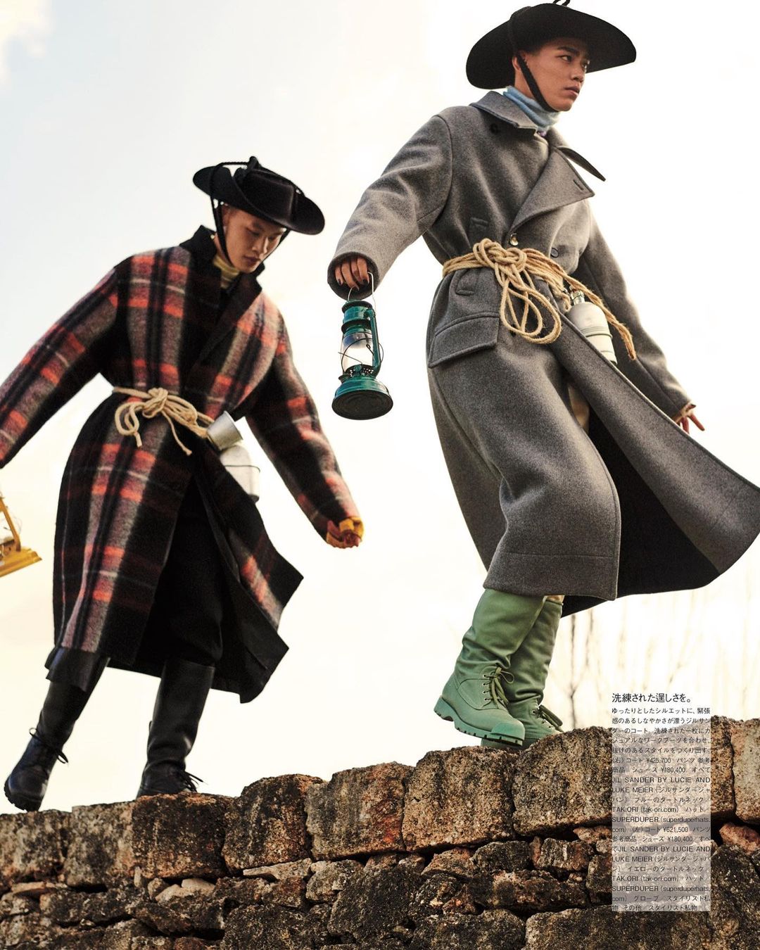 Two adult models pose in long coats, wide-brimmed hats and boots on a stone wall; one carries a lantern against a bright, outdoor background.