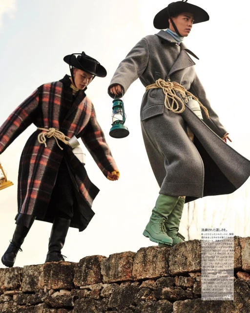 Two adult models pose in long coats, wide-brimmed hats and boots on a stone wall; one carries a lantern against a bright, outdoor background.