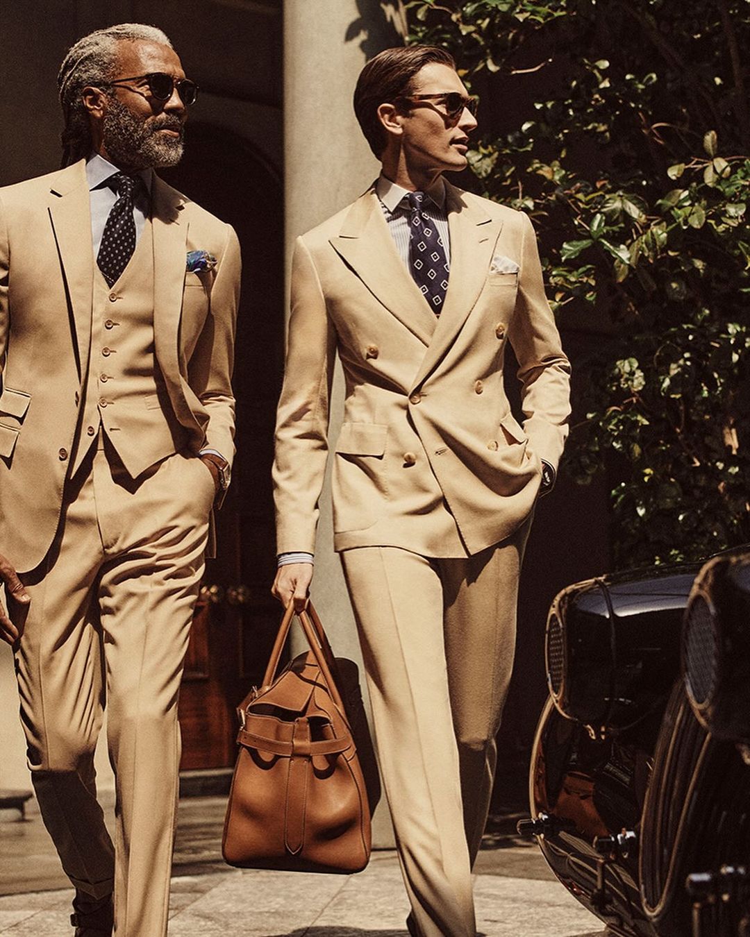 Two stylish men in beige suits walk outdoors, one carrying a duffel bag, near a classic car, showcasing men's fashion.