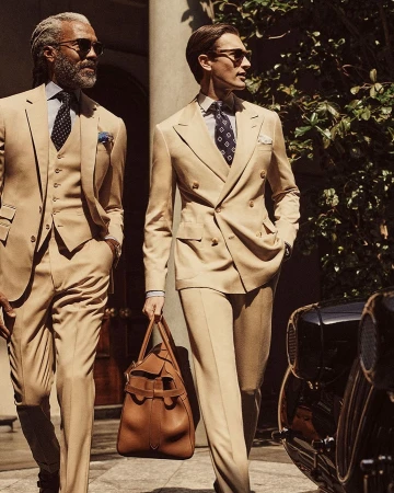 Two stylish men in beige suits walk outdoors, one carrying a duffel bag, near a classic car, showcasing men's fashion.