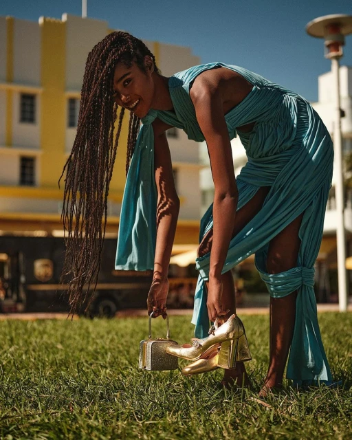 A woman in a blue dress poses on a grassy lawn with golden high heels and a small purse, with a building in the background.