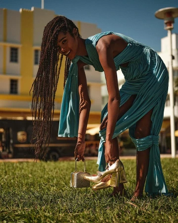A woman in a blue dress poses on a grassy lawn with golden high heels and a small purse, with a building in the background.