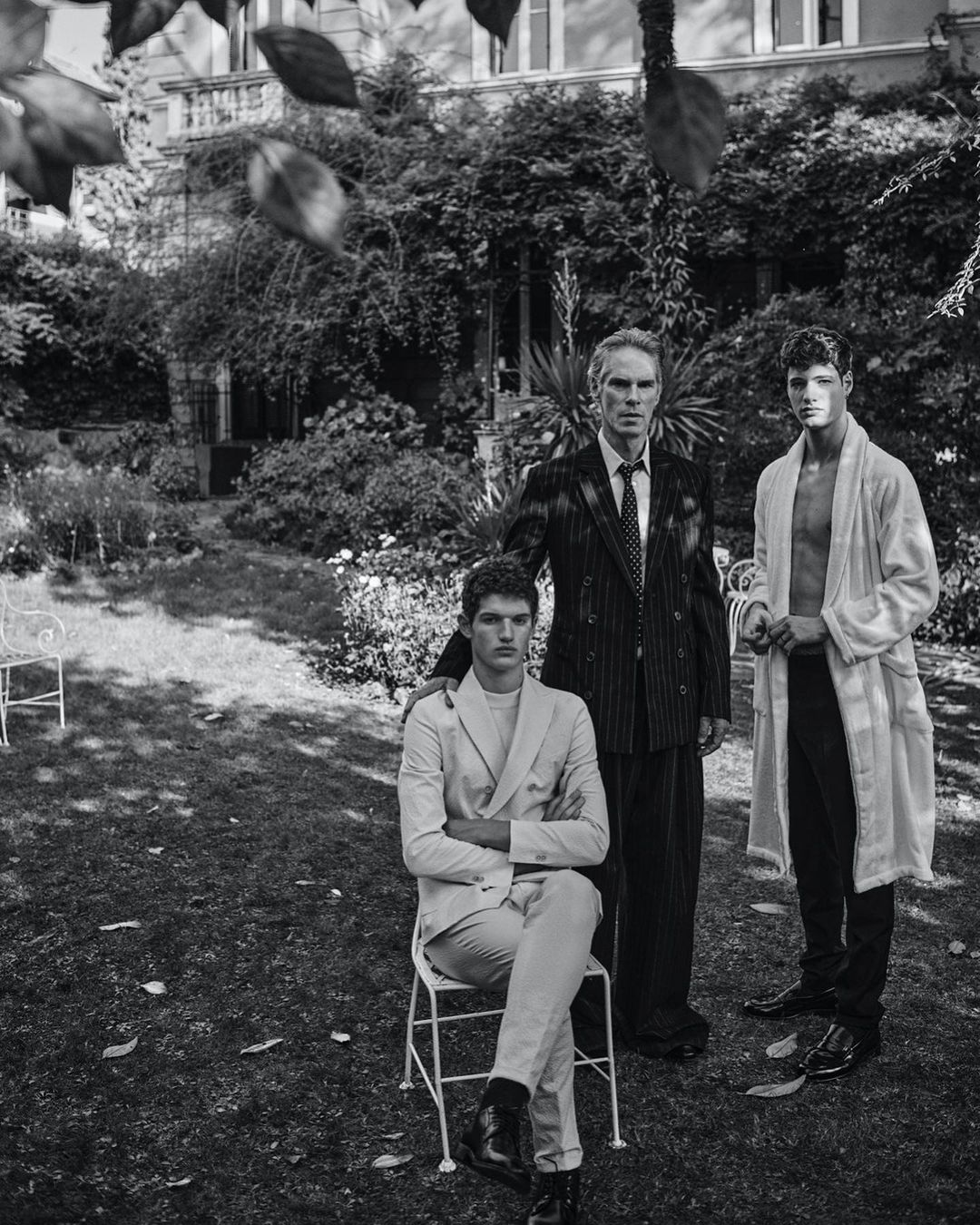 Black and white outdoor shot of three men of varying ages standing together in a grassy yard in front of a vine-covered house.