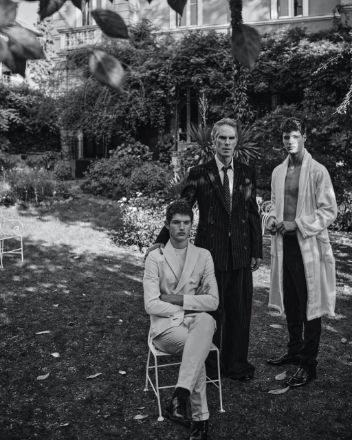 Black and white outdoor shot of three men of varying ages standing together in a grassy yard in front of a vine-covered house.