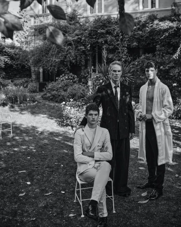 Black and white outdoor shot of three men of varying ages standing together in a grassy yard in front of a vine-covered house.